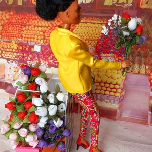 May include: A doll wearing a yellow blazer and red patterned pants, holding a bouquet of flowers. A display of flowers in a black and white checkered box is in the foreground. The background shows a fruit stand with signs that read "Florida Fresh" and "California".