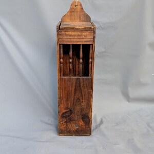 May include: A tall, rectangular wall-mounted wooden box with a dark brown stained finish. The upper section features a series of vertical spindles, while the top has a shaped, arched backboard with a small hole for hanging on a wall.