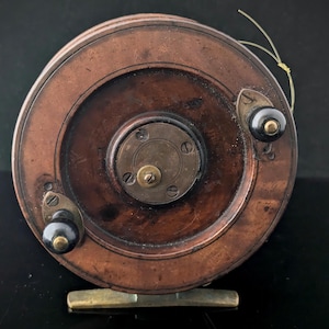 Könnte beinhalten: Eine Vintage-Angelrolle mit einem kreisförmigen, kupferfarbenen Gehäuse. Sie verfügt über schwarze Knöpfe, Messingakzente und einen Messinggriff. Eine dünne, gelbe Angelschnur ist sichtbar. Die Rolle ist zum Angeln bestimmt.