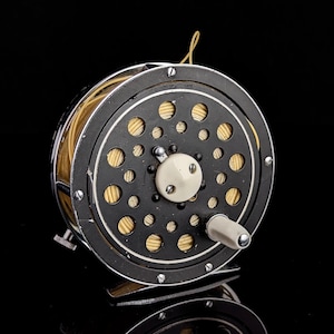 Pode incluir: Um molinete de pesca circular com uma estrutura de metal preto com uma série de recortes circulares, um botão central branco e uma alça branca. O molinete contém um carretel de linha de pesca bege claro visível através da placa lateral.