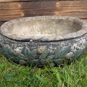 Op de afbeelding: Een ronde, verweerde stenen plantenbak met een donkergrijze en groene afwerking, voorzien van een reliëfpatroon van gebeeldhouwde bladeren en eikels rondom de buitenkant. De bak heeft een dikke, afgeronde rand en een diepe, open binnenkant.