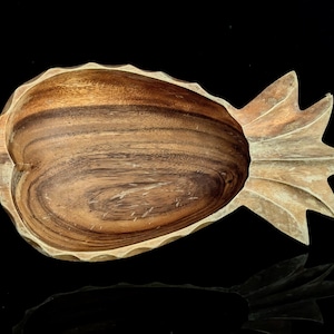 Pode incluir: Uma tigela de madeira esculpida em forma de abacaxi, apresentando uma bacia profunda e arredondada com padrões de veios de madeira natural visíveis em tons de castanho e bege, e uma secção de folhas pontiagudas e texturizadas num lado.
