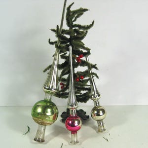 Può includere: Tre puntali per albero di Natale in vetro vintage con estremità appuntite allungate e basi sferiche. Un puntale presenta una sfera verde, uno una rosa e uno una argentata, tutti con finiture metalliche e steli in vetro trasparente.