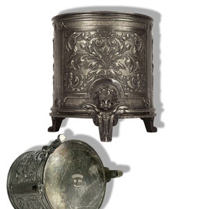 Pode incluir: Uma caixa de metal prateada com intrincados desenhos florais e uma cabeça de querubim na frente. A caixa tem três pernas e uma tampa com dobradiças.