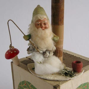 Może przedstawiać: Winietkowa ozdoba choinkowa przedstawiająca małą figurkę gnoma z białą brodą i zielonym kapeluszem, trzymającego wędkę z czerwono-białym grzybkiem na końcu. Gnom siedzi w śnieżnej scenerii z podstawą z szyszki i białą kulką z waty symbolizującą śnieg. Ozdoba jest umieszczona na drewnianej podstawie z zielonym wzorem ostrokrzewu.