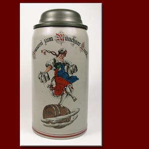 May include: A stoneware beer stein with a hinged lid. The stein features a colourful illustration of a woman in traditional Bavarian attire balancing on a barrel, holding beer steins. The text "Brauerei zum Münchner Kindl" is arched across the top.