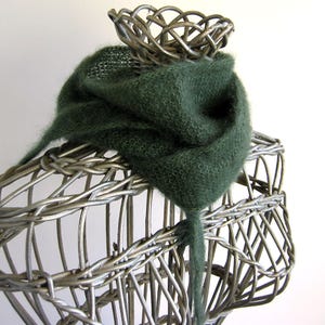 Może przedstawiać: Ciemnozielony, dziergany szalik o miękkiej, puszystej fakturze, narzucony na drucianą formę w kształcie kosza. Długi koniec szalika zwisa. Mały druciany kosz znajduje się na górze. Tło jest białe.