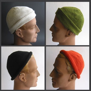 Könnte beinhalten: Vier gestrickte Mützen in verschiedenen Farben. Die Mützen haben alle den gleichen Stil, mit einem umgeschlagenen Rand und einer lockeren Passform. Die Farben sind weiß, grün, schwarz und orange.