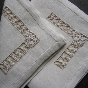 Könnte beinhalten: Zwei cremefarbene Leinenservietten mit zartem weißen Spitzenbesatz. Der Spitzenbesatz ist in einem geometrischen Muster.