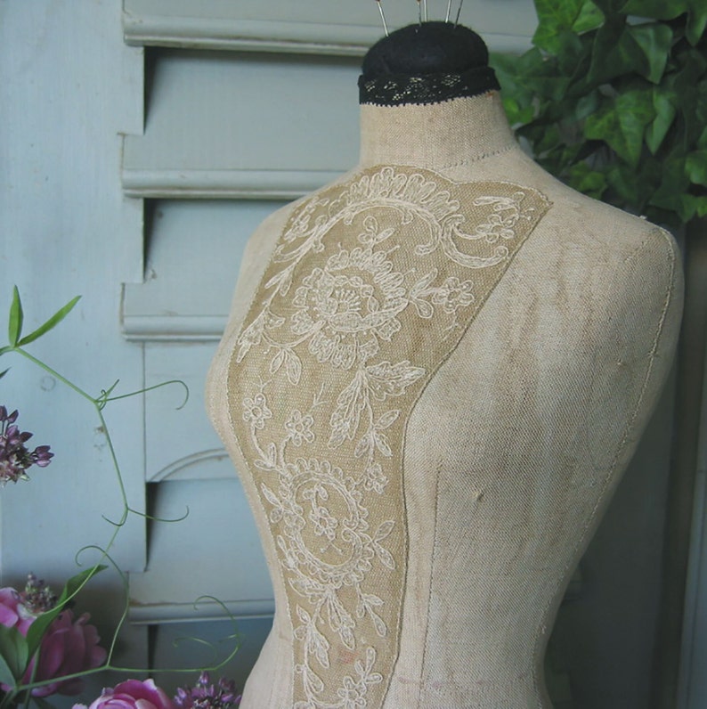 May include: A vintage fabric dress form with a delicate white lace design on the bodice. The form is made of a beige fabric and has a simple, classic design.