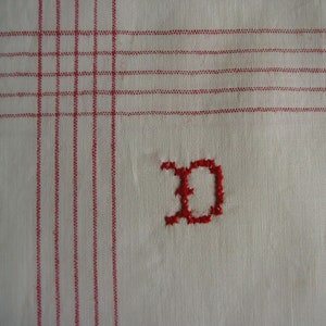 Pode incluir: Tecido branco com linhas bordadas vermelhas e uma letra "D" bordada vermelha no centro.