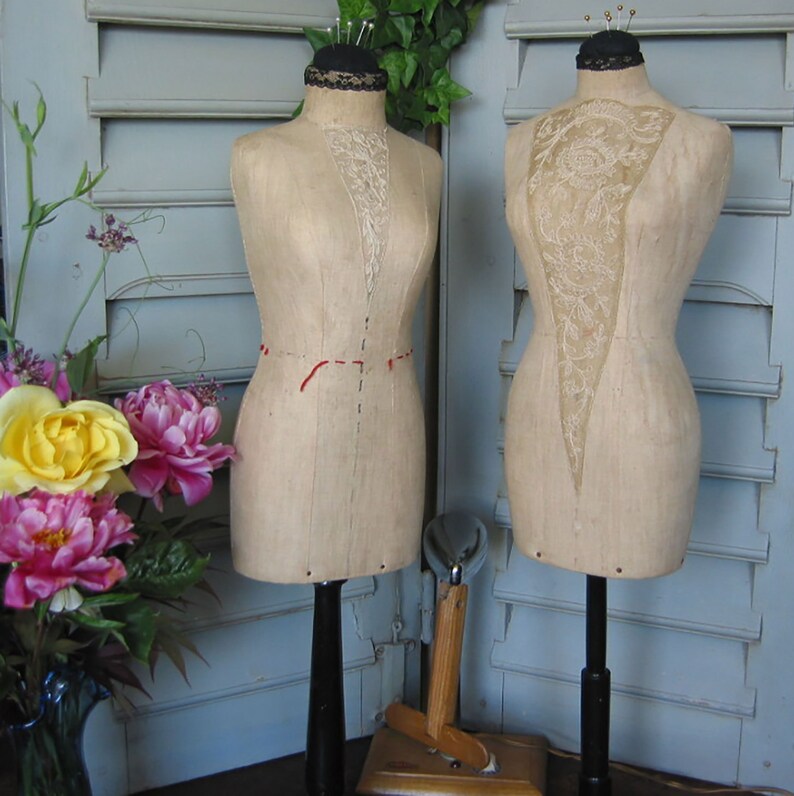 May include: Two vintage dress forms with fabric coverings on stands. The dress forms are made of a light brown material and have a lace overlay. The stands are black and the dress forms are against a white shuttered background.