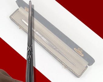 May include: A silver-colored wand with intricate detailing and a decorative design. The wand is displayed in a black case with a gold-colored trim. The wand has a sleek, elongated shape.