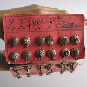 Könnte beinhalten: Vintage Set von "The Reliable Lever Studs" auf einer roten Displaykarte. Die Karte zeigt den Text "English Make" und eine Schiffsillustration. Die Manschettenknöpfe sind metallisch, mit verschiedenen dekorativen Designs.