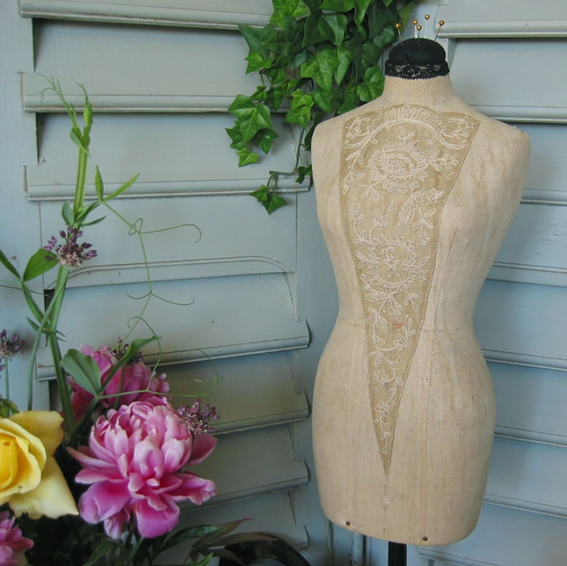 May include: A vintage dress form with a lace overlay, standing on a wooden base. The dress form is made of a beige fabric and has a delicate, floral design. The lace overlay is white and has a delicate, floral design. The dress form is standing on a wooden base.