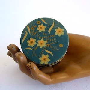 Peut inclure: Un ancien récipient rond bleu sarcelle avec un motif floral de jonquilles jaunes et des accents dorés. Le récipient porte l'inscription "Le Narcisse Bleu" et "Mury Paris France".