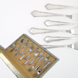 Könnte beinhalten: Ein Set aus Silberbesteck und Servierutensilien. Die Gabeln befinden sich in einer beigefarbenen Box mit hellgrüner Umrandung. Mehrere Gabeln sind außerhalb der Box angeordnet. Die Utensilien haben einen dekorativen Rand.