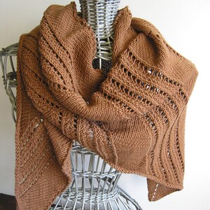 Könnte beinhalten: Ein braunes gestricktes Tuch mit einem Spitzenmuster. Das Tuch ist über eine weiße Weidenpuppe gelegt.