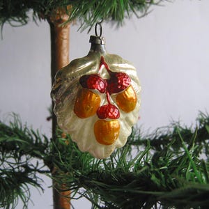 Puede incluir: Adorno navideño vintage con forma de hoja, con bayas rojas y bellotas doradas. El adorno de cristal tiene una tapa plateada para colgarlo y está colgado de un pequeño árbol de Navidad artificial.