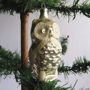 Puede incluir: Adorno navideño vintage con forma de búho. El búho de cristal plateado tiene detalles blancos y brillantes. Cuelga de un lazo de metal plateado entre ramas verdes de árbol de Navidad.