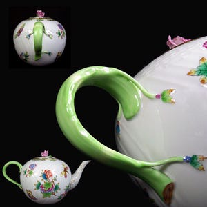 Könnte beinhalten: Eine weiße Teekanne mit einem grünen Henkel und einem Blumenmuster. Die Teekanne hat eine rosa Blume auf dem Deckel und ist mit grünen Blättern und bunten Blumen verziert.