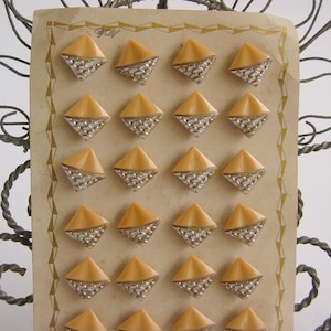 May include: A selection of vintage diamond-shaped buttons. Each button has a light orange, pyramid-shaped top and a silver-coloured, textured base. The buttons are arranged on a cream-coloured card with a decorative border.