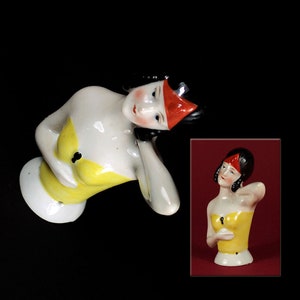 Könnte beinhalten: Eine Porzellanfigur einer Frau mit schwarzen Haaren und einem roten Stirnband. Sie trägt ein gelbes Kleid und hat ein Schlüsselloch auf der Brust. Die Figur ist in einer liegenden Pose.