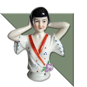 Könnte beinhalten: Porzellanfigur einer Frau mit schwarzen Haaren, die einen weißen Kimono mit roten Verzierungen und blauen Punkten trägt. Die Figur hat die Hände an ihrem Kopf. Eine lila Blume befindet sich auf dem Kimono.