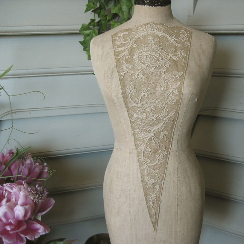 May include: A vintage, off-white fabric dress form with a delicate lace design on the front. The lace is a light cream color and features a floral pattern.