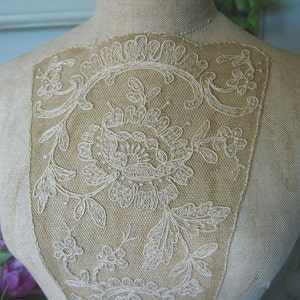 May include: A close-up of a white lace design on a beige fabric mannequin. The lace is a delicate floral pattern with intricate details.