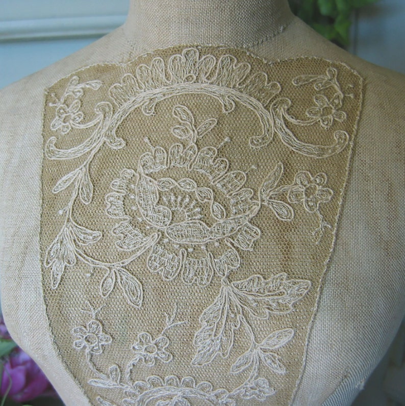 May include: A close-up of a white lace design on a beige fabric mannequin. The lace is a delicate floral pattern with intricate details.