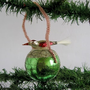 May include: A retro Christmas tree decoration featuring a bird atop a green bauble. The bird has a silver body, red details, and a white tail. It hangs from a pink cord, set against a backdrop of green fir branches.