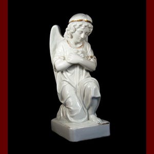 May include: A white porcelain angel figurine kneeling with hands clasped in prayer. The angel has a gold halo and gold accents on the wrists and neckline. The figurine is on a rectangular base.
