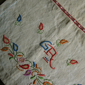 May include: A white linen tablecloth with embroidered floral designs in shades of blue, red, orange, green, and purple. The embroidery includes a stylised letter 'H' in red and blue.