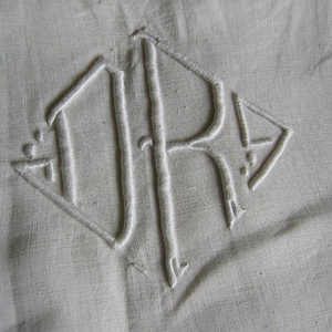 Könnte beinhalten: Weiß gesticktes Monogramm der Buchstaben "JR" in Diamantform auf einem weißen Leinenstoff.