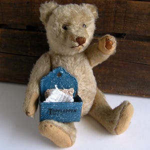 May include: A vintage teddy bear with light brown fur and a stitched nose and mouth. The bear holds a small, blue metal container with the word "TOPFLAPPEN" and a white and tan item inside.