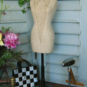 May include: A vintage dress form with a lace bodice and a beige fabric body. The dress form is on a black stand with a round base. The dress form is in front of a light blue wall with white shutters.