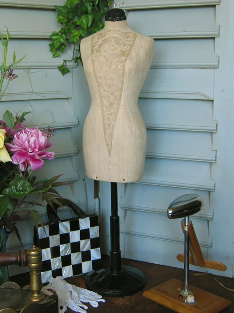 May include: A vintage dress form with a lace bodice and a beige fabric body. The dress form is on a black stand with a round base. The dress form is in front of a light blue wall with white shutters.