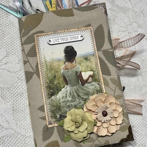 May include: A handmade journal with a fabric cover featuring a floral pattern and a picture of a woman reading. The journal has decorative paper flowers and ribbons. The text "LIVE YOUR STORY" is visible on the cover.