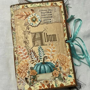 May include: A handmade scrapbook with an autumn theme. The cover features a teal pumpkin, autumn leaves, and flowers. The title of the book is "Album" and the text "Autumn is... Hayrides, Bonfires, Pumpkins, Apples, Leaves" is on the cover.