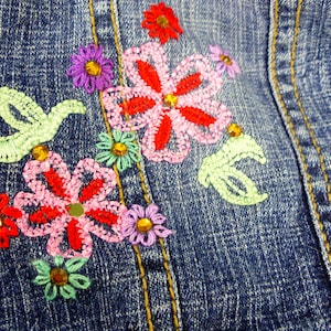 May include: A close-up of a denim jacket with embroidered flowers in shades of pink, red, green, and purple. The flowers are detailed with sequins and beads.