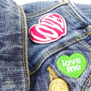 May include: A blue denim jacket with two heart-shaped patches. The top patch is pink with the word "LOVE" written inside. The bottom patch is green with the words "LOVE ME" written inside.
