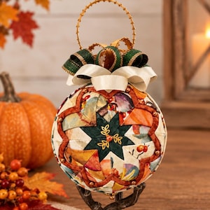 May include: A spherical fabric ornament featuring a quilted star pattern in shades of dark green, orange, and cream. The ornament is topped with a layered bow of green and cream ribbons and a gold wire loop. A small gold leaf charm is centered.
