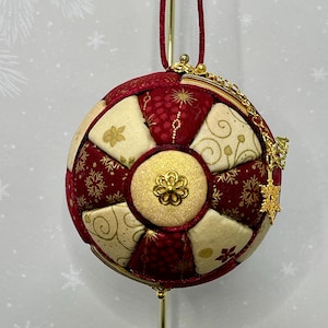 May include: A round Christmas ornament with a red and cream patchwork design. The ornament features gold snowflake and floral accents, a gold clasp, and a gold chain with a snowflake charm. A red cord is attached for hanging.