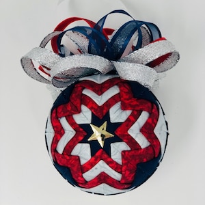 May include: A red, white and blue quilted Christmas bauble with a gold star in the centre. The bauble is topped with a silver and blue ribbon.