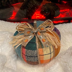 May include: A plaid fabric pumpkin with a burlap bow and a straw accent. The pumpkin is decorated with a ribbon featuring a pattern of small pumpkins and candy corn.