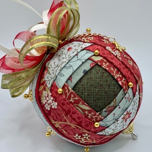 May include: A red, green, and white fabric Christmas ornament with a gold chain and gold beads. The ornament is decorated with a red and white ribbon.