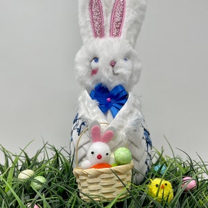 May include: A white plush Easter bunny with pink glitter-lined ears and a blue bow tie. The bunny holds a small basket with a white bunny, a carrot, and colorful eggs. The scene is set in artificial grass with more Easter eggs and a yellow chick.