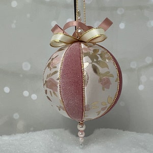 May include: A pink and white Christmas ornament with a floral pattern and gold accents. The ornament is decorated with a pink satin ribbon bow and a pearl string.