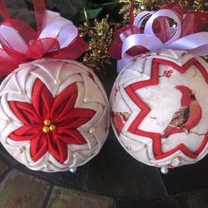May include: Two handmade Christmas ornaments, one with a red poinsettia design and the other with a cardinal bird design. Both ornaments are white with red accents and have a white and red bow.